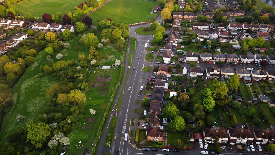 An aerial view of a suburban residential area featuring a main road with moving cars, flanked by neatly maintained gardens and lawns. To the left, a large green space divided into garden beds and gras